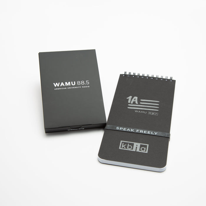 Black custom-branded spiral notebook with "1A WAMU NPR" and "SPEAK FREELY" printed on the cover, next to a matching black box labeled "WAMU 88.5 American University Radio.