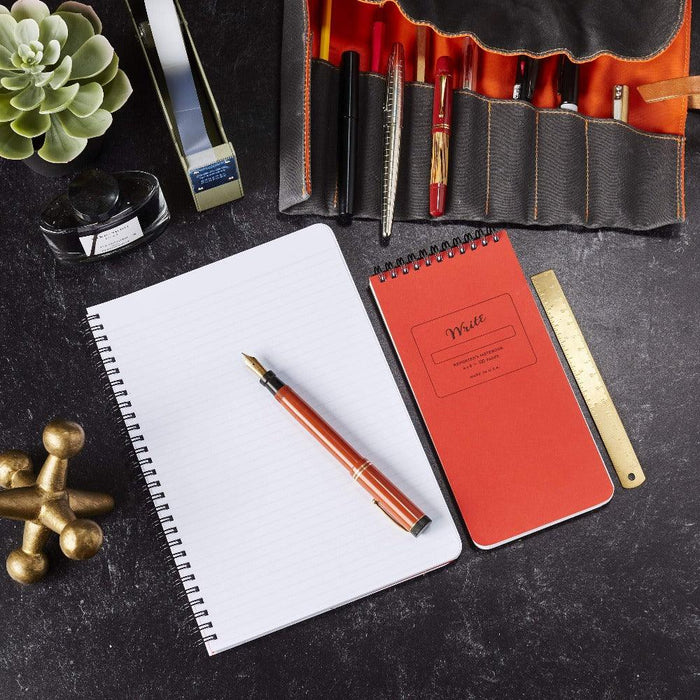 Lined notebook on a desk with a high-end fountain pen on top, surrounded by a open pen case with multiple pens in the slots.