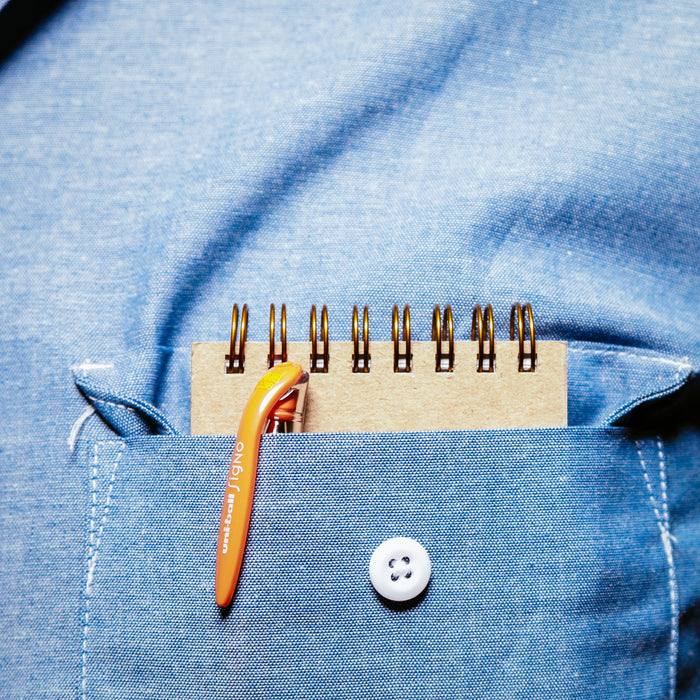 Close-up of a denim shirt pocket with a 'Write' Pocket Ledger peeking out, accompanied by an orange pen clip. The light blue fabric has a prominent stitch line and a white button.