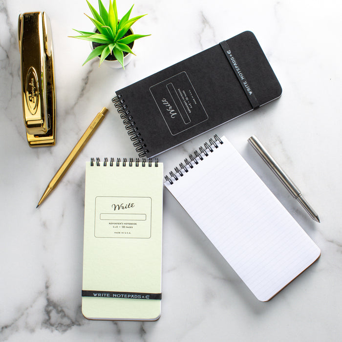 Flat lay of office essentials: a gold stapler, mint-green 'Write' Pocket Ledger, dark gray 'Write' Pocket Ledger with embossed lettering, open blank white spiral notebook, golden pencil, and silver pen, all on a marble background with a small green succulent plant.