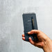 Hand holding the 'Write' Pocket Ledger with a sleek gold-trimmed pen attached. The black notebook's cover reads 'POCKET LEDGER' and has a spiral top-binding, against a textured gray background.